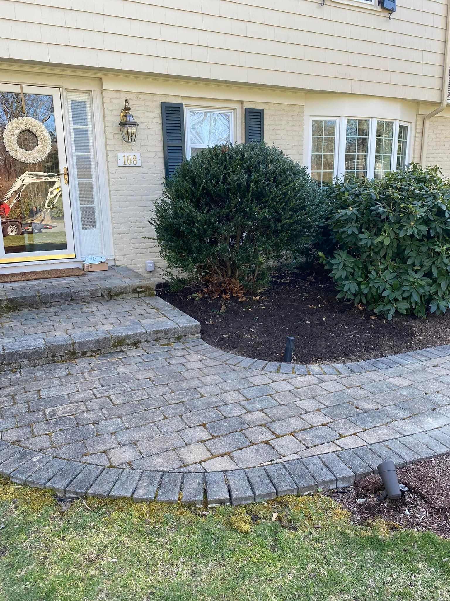 Front entrance of a house with stone steps, curved paver walkway, shrubs, and a wreath on the door
