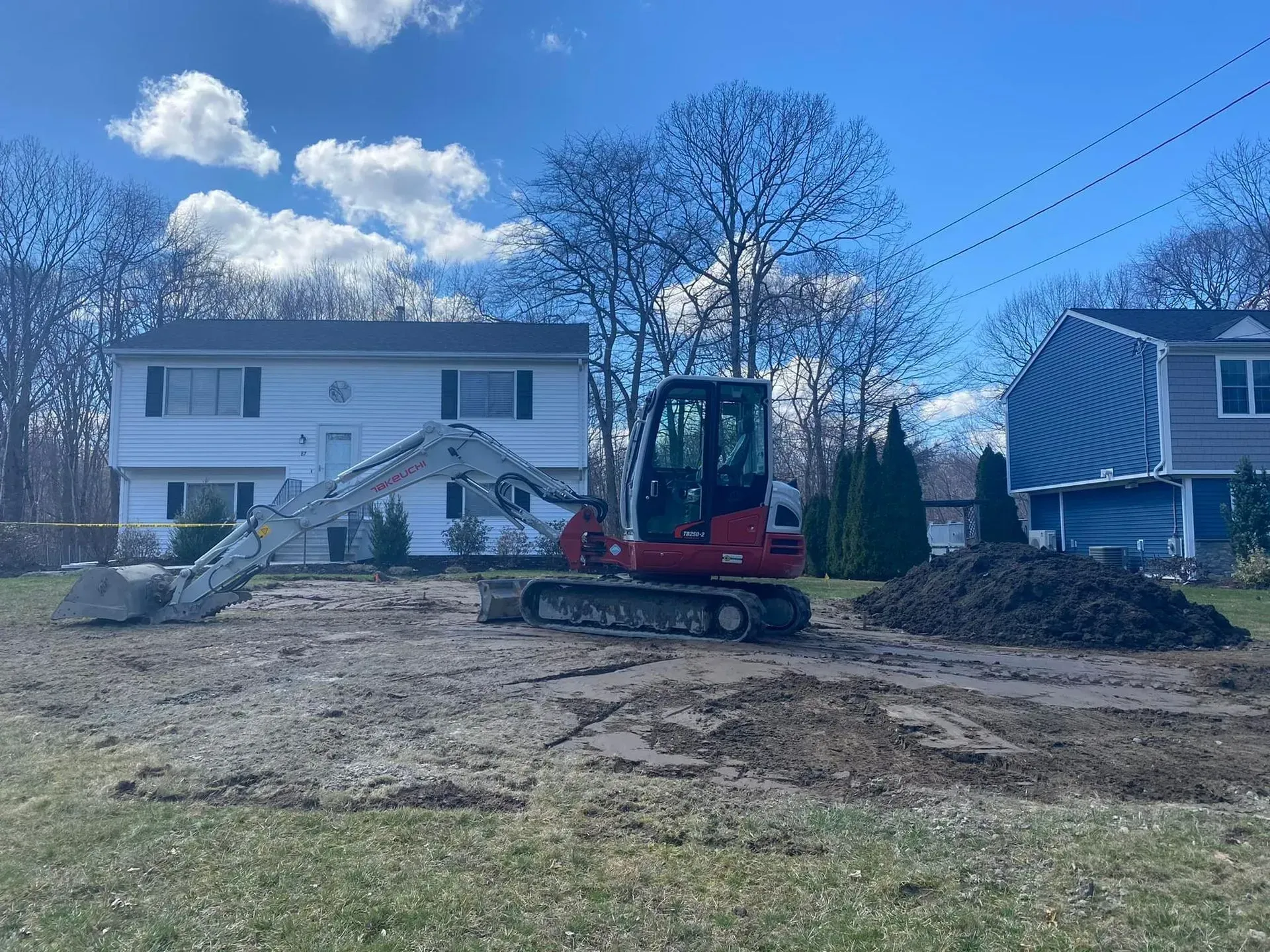 Red excavator digging dirt in front of a white two-story house on a cloudy day