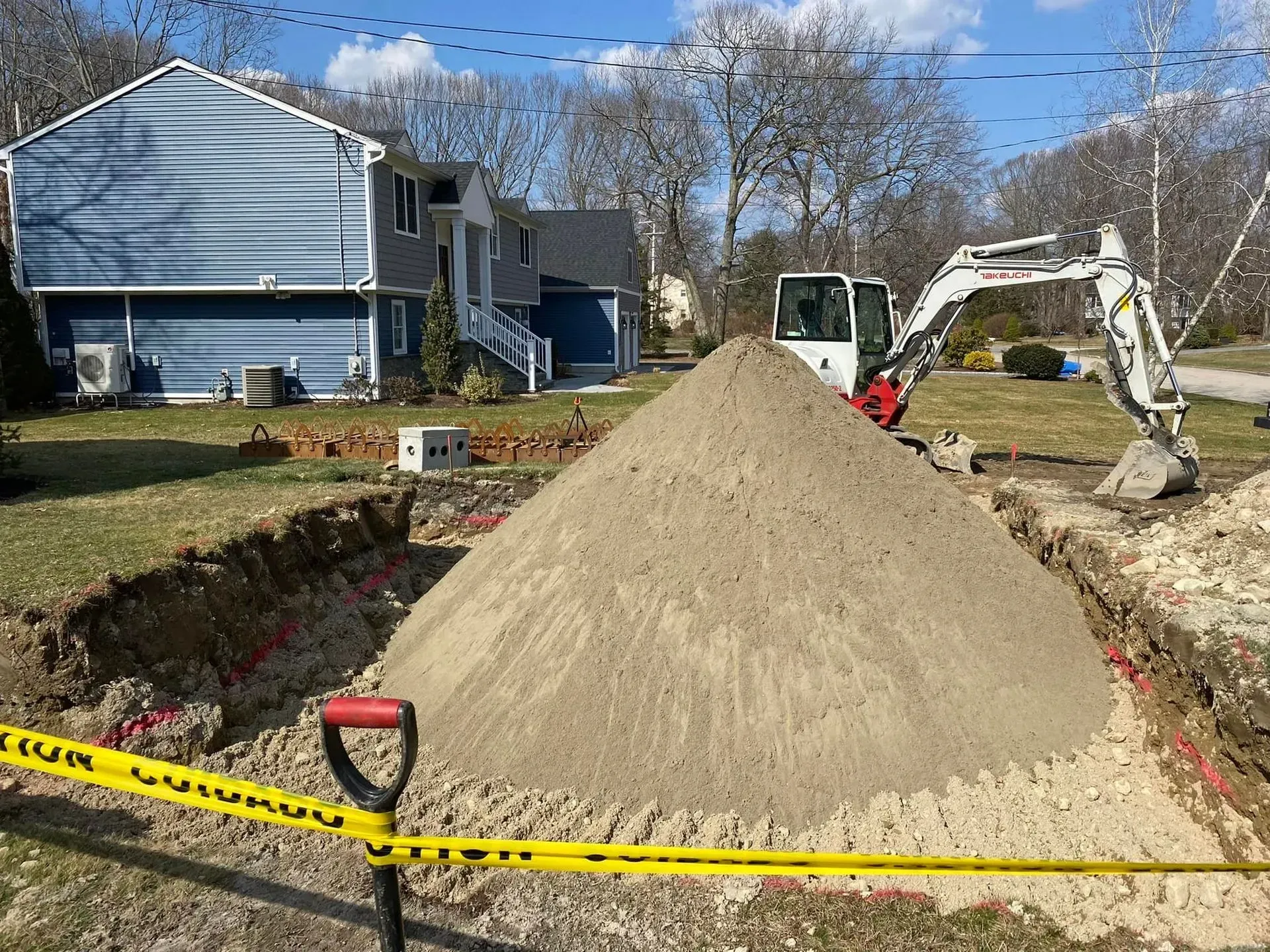 Sand pile and excavator in a trench outside a blue house, with caution tape across the yard