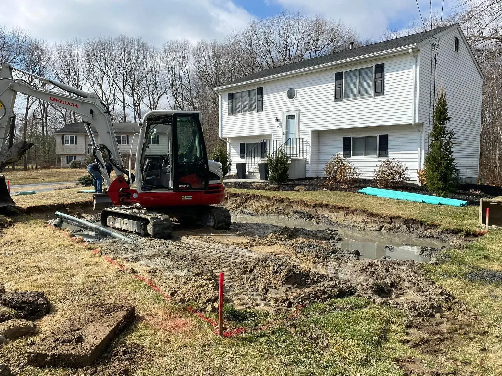 Small excavator digging a yard trench beside a white house on a muddy construction site