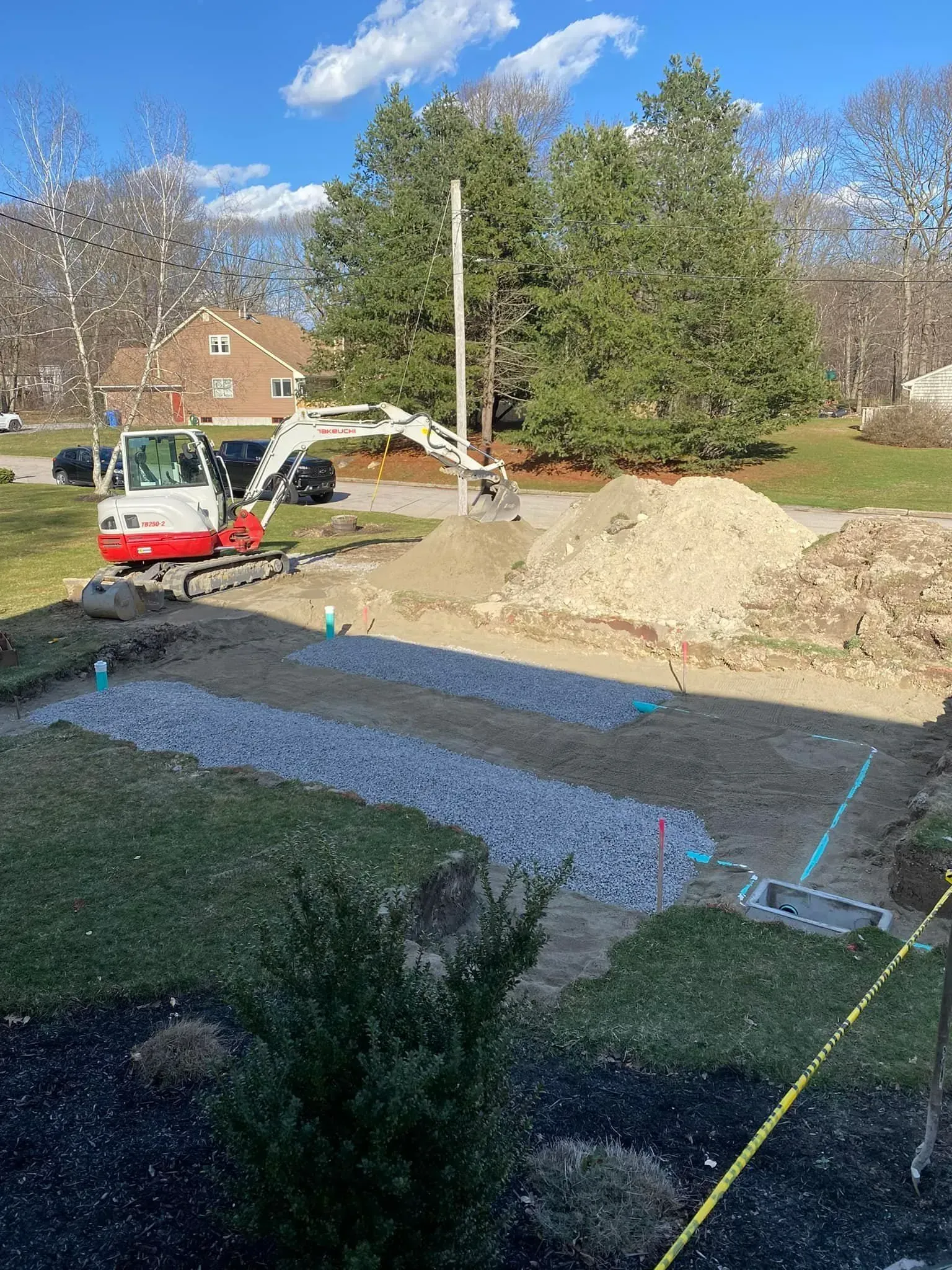 Excavator grading a backyard beside a large dirt pile and marked blue utility lines on a sunny day
