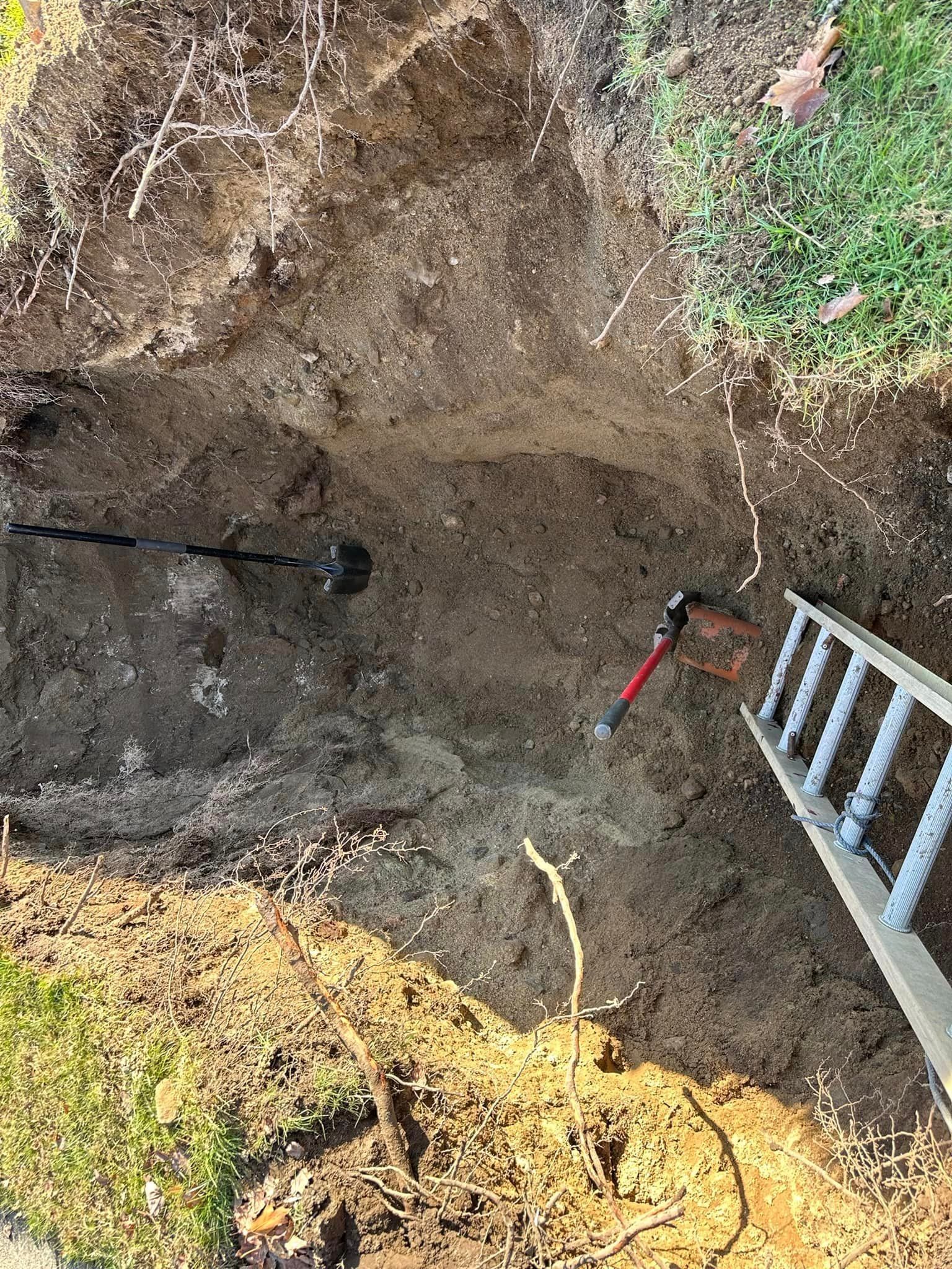 Shallow dirt pit with a ladder on the right and a shovel and pickaxe inside a grassy yard