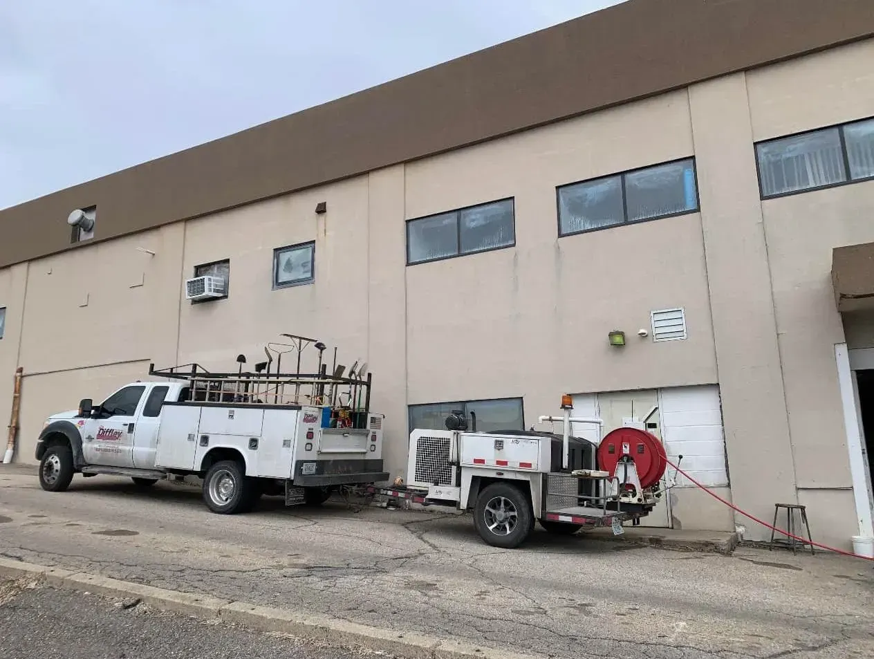 White utility truck and trailer with equipment parked beside a beige industrial building.