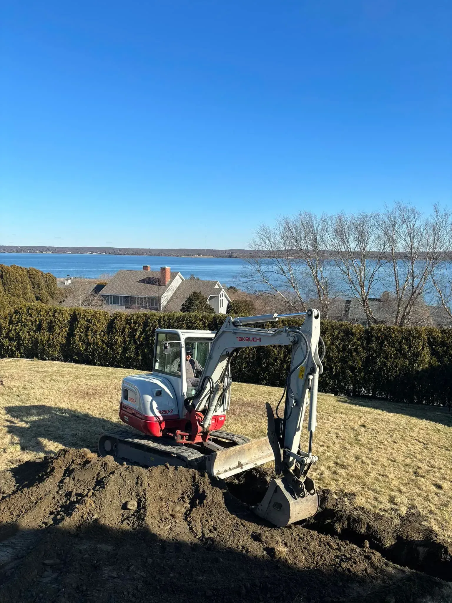 Small excavator digging a trench beside a waterfront yard on a clear day.