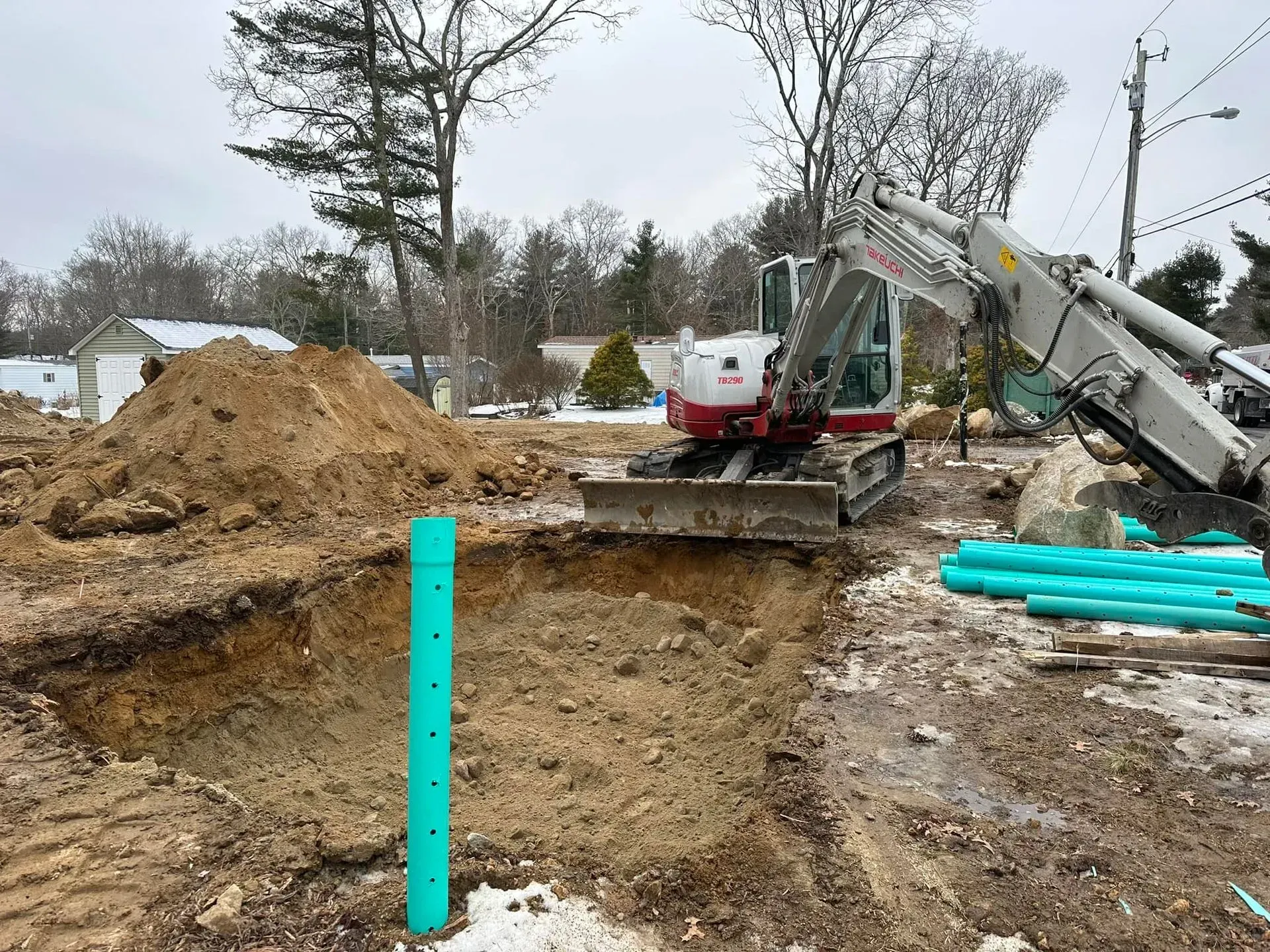 Excavator digging a large hole at a muddy construction site with piles of dirt and turquoise pipes.