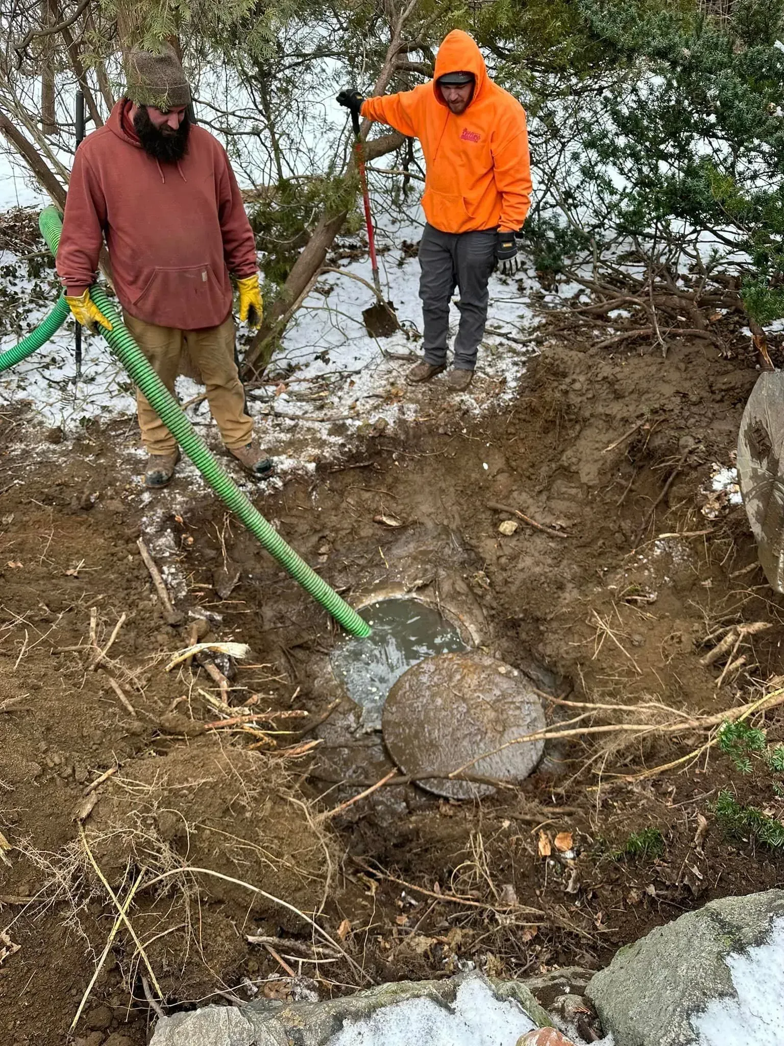 Two people in winter coats inspect a muddy creek bank with a green hose pumping water.