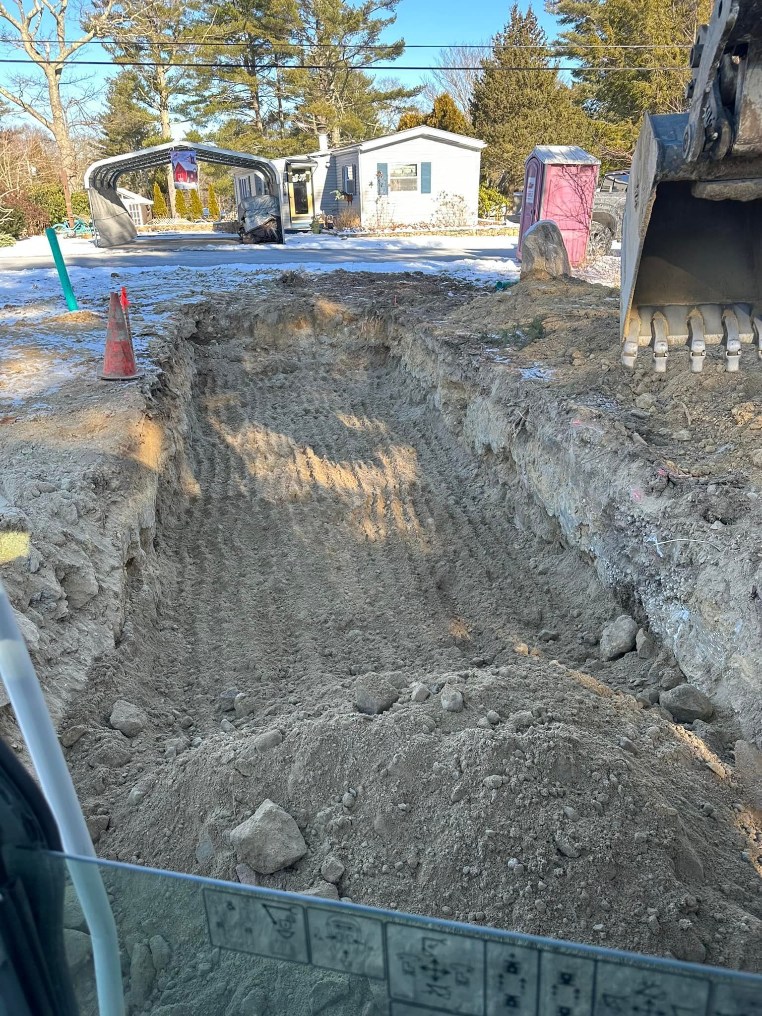 Construction site with a deep gravel trench, orange cone, and excavator bucket on the right
