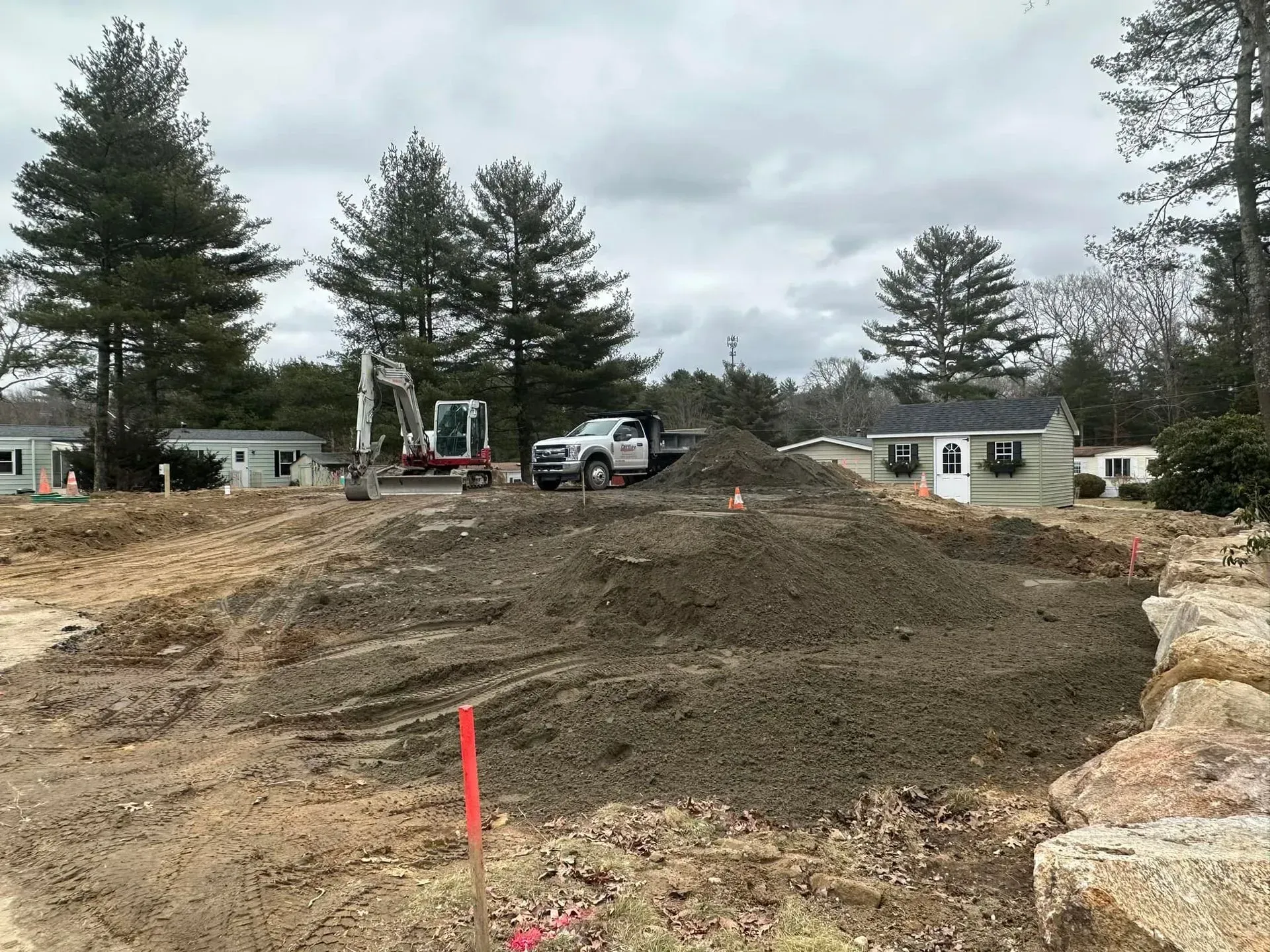 Construction site with excavator and truck shaping a dirt mound between tall trees and small buildings