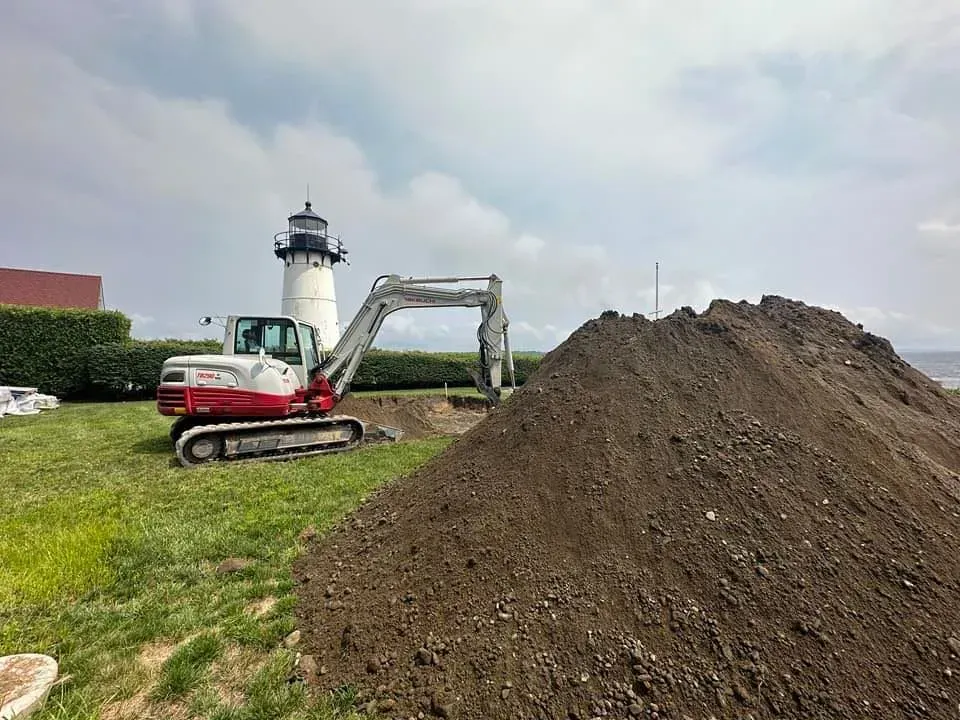 Excavator beside a large dirt pile near a lighthouse on a grassy coast