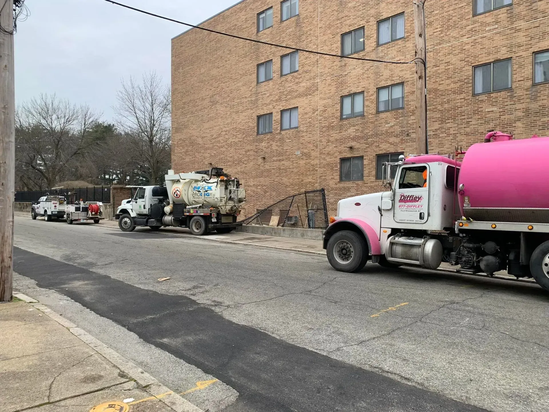 Trash trucks lined along a street beside a brick apartment building, including a pink tanker truck in front.