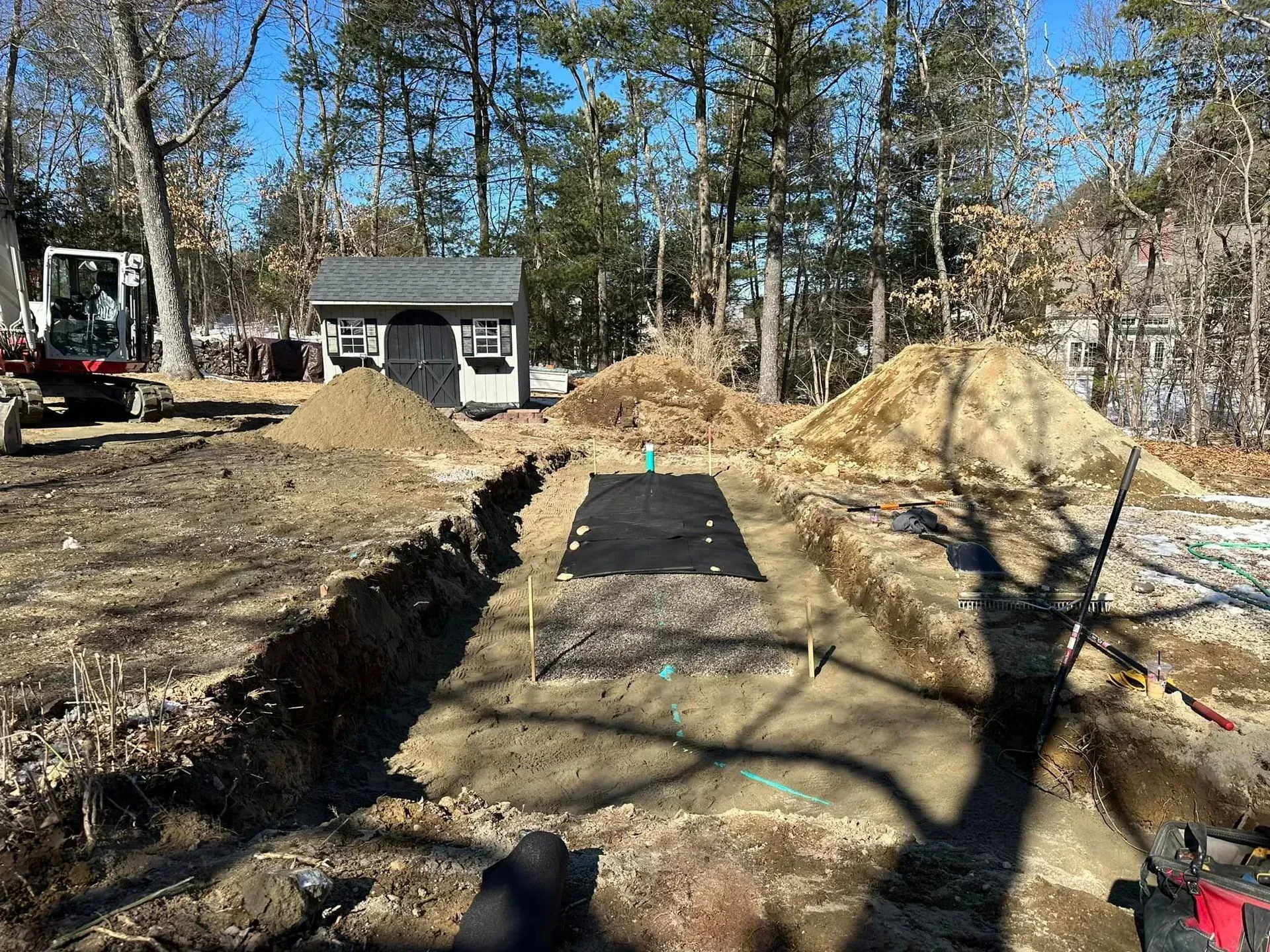 Trench with black ground cover in a wooded yard, flanked by dirt piles and a small house in the background