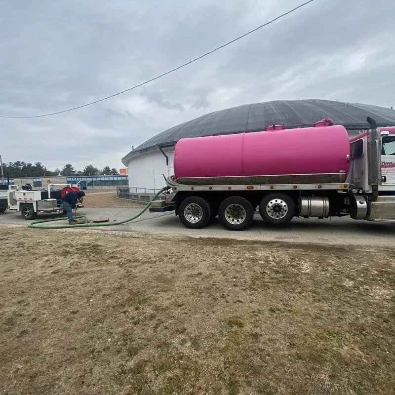Pink water tanker truck parked beside a white silo on a farm, with a hose running to a smaller truck.