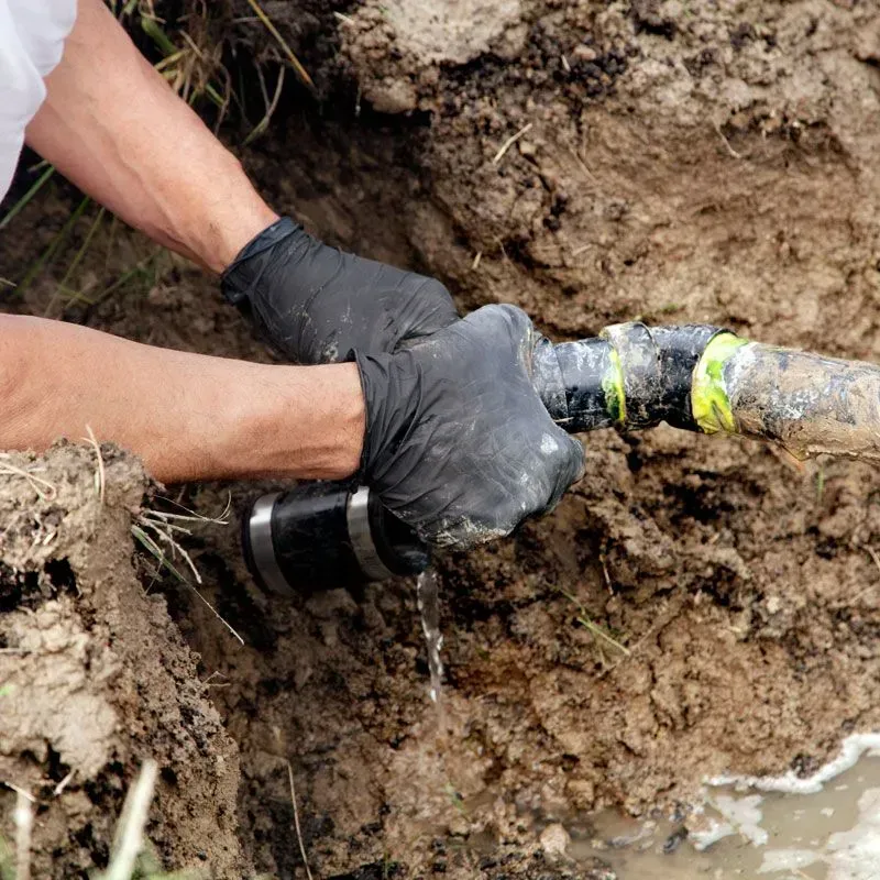 Gloved hands connecting a black pipe fitting in a muddy trench outdoors