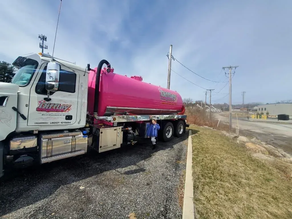 Pink tanker truck parked on a gravel roadside beside a highway on a cloudy day