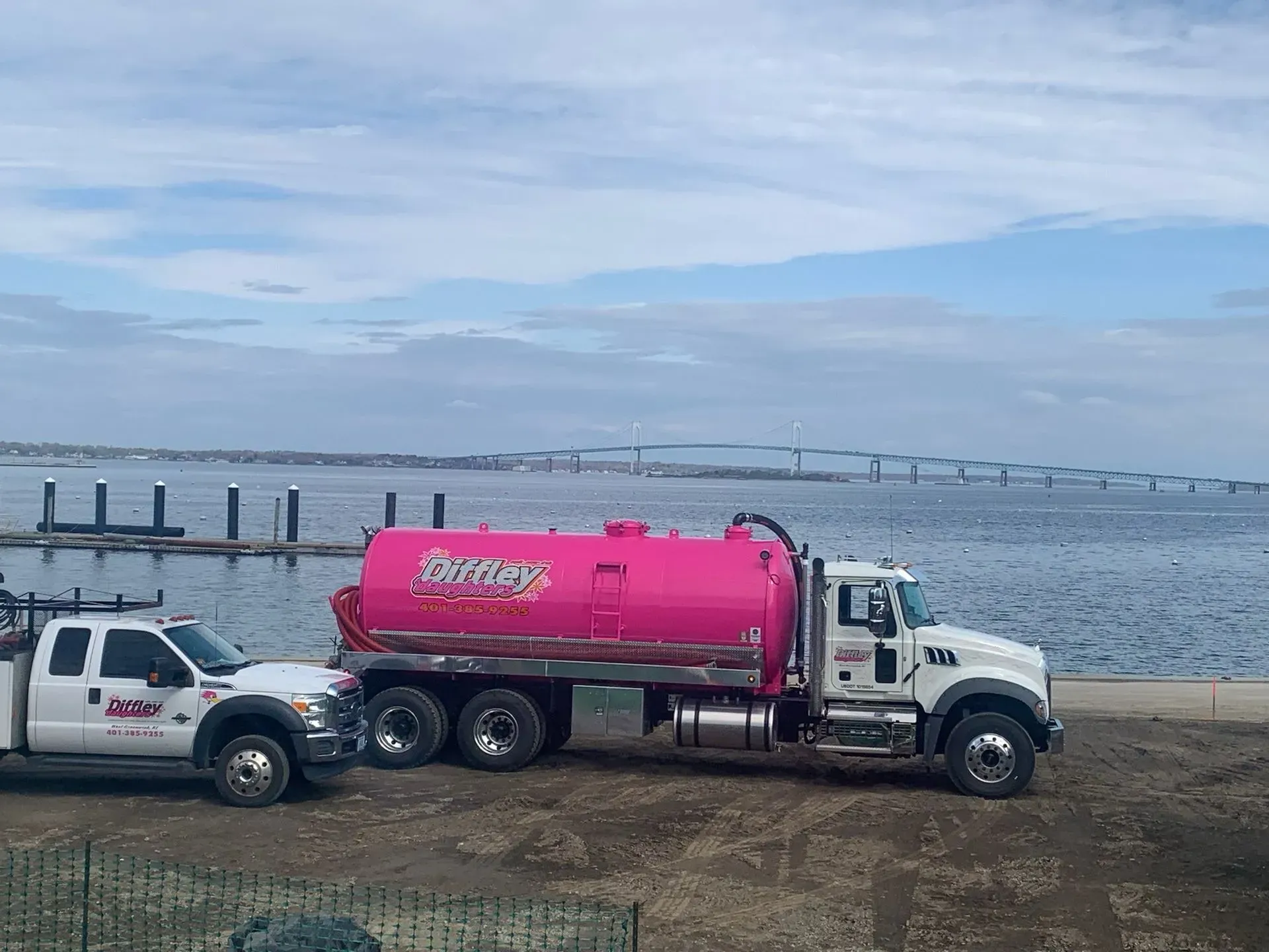Pink tanker truck and white pickup parked by a waterfront under a cloudy sky