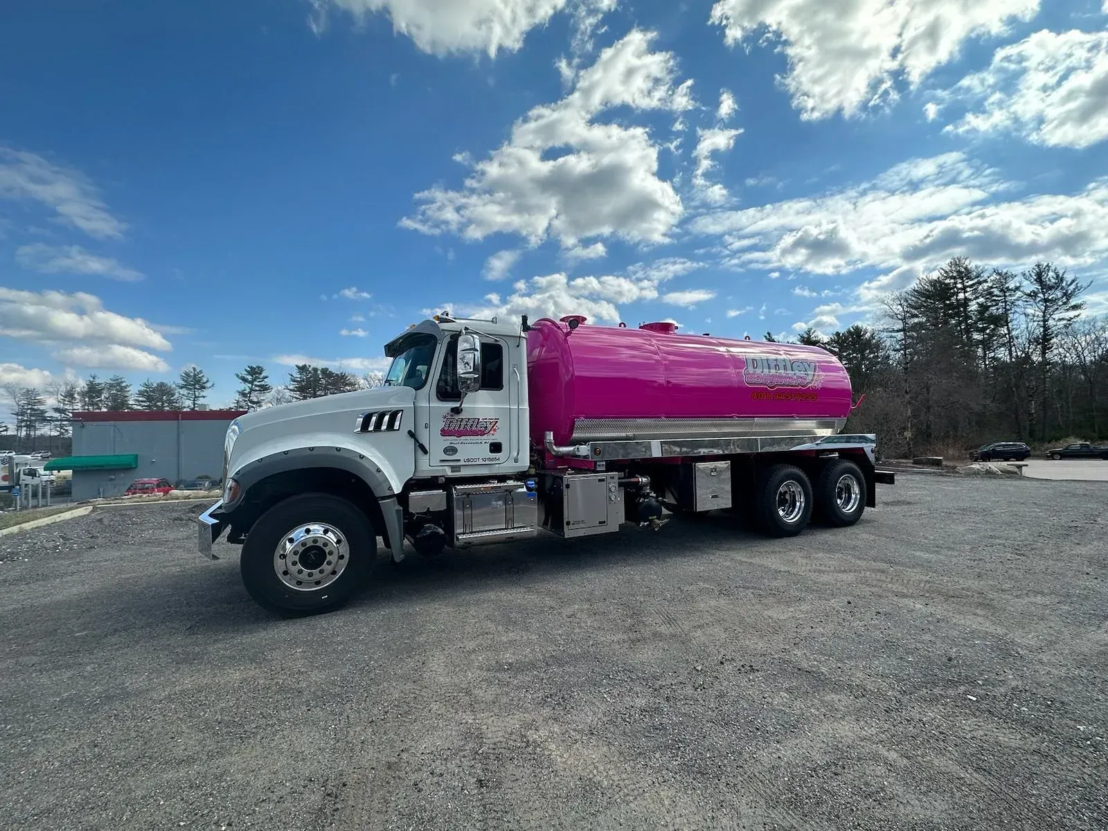 White tanker truck with a bright pink tank parked on gravel under a cloudy blue sky