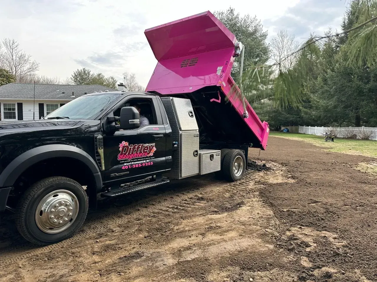 Black dump truck with raised pink bed parked on a dirt lot near a house and trees.