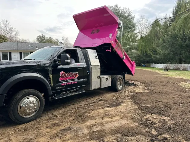 Black dump truck with bright pink raised bed parked on a dirt lot beside a house and trees.