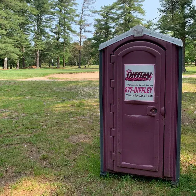 Maroon portable toilet in a grassy park with trees in the background