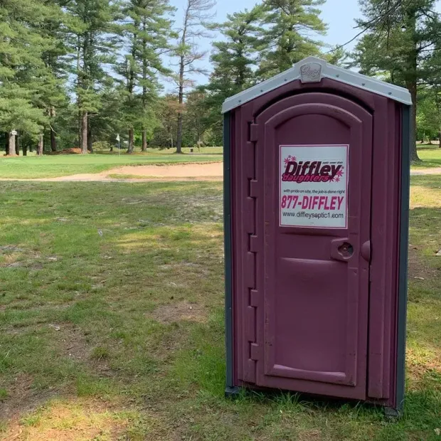Purple portable toilet with sign in a grassy park area near trees