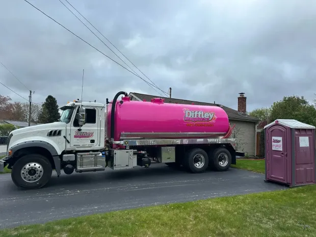 Pink tanker truck parked beside a driveway near a purple portable toilet under cloudy skies
