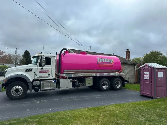 Pink tanker truck parked beside a driveway near a purple portable toilet under cloudy skies