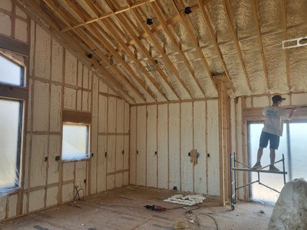 A worker stands on a scaffold inside an unfinished room with walls and a vaulted ceiling covered in spray foam insulation.