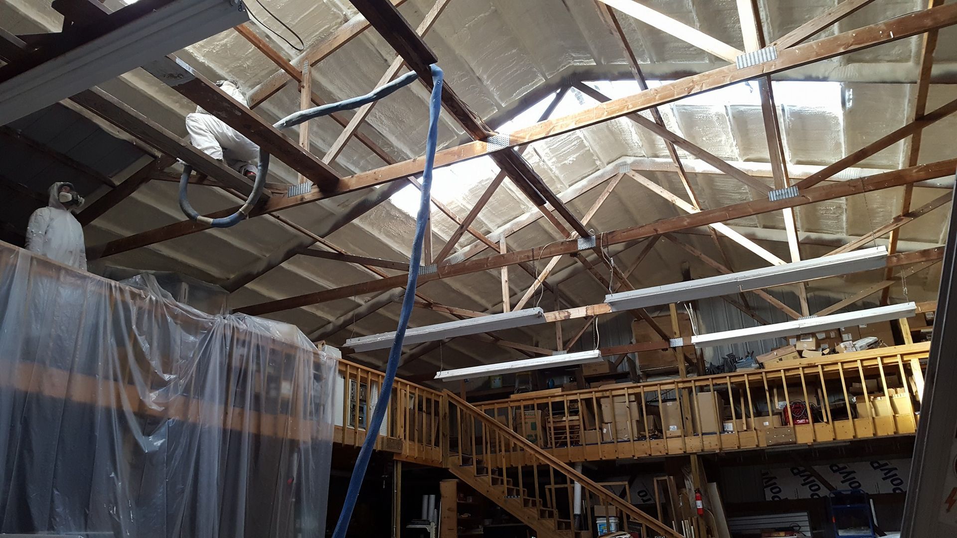 An interior view of a workshop with wooden rafters, skylights, hanging lights, and a staircase leading to a loft.