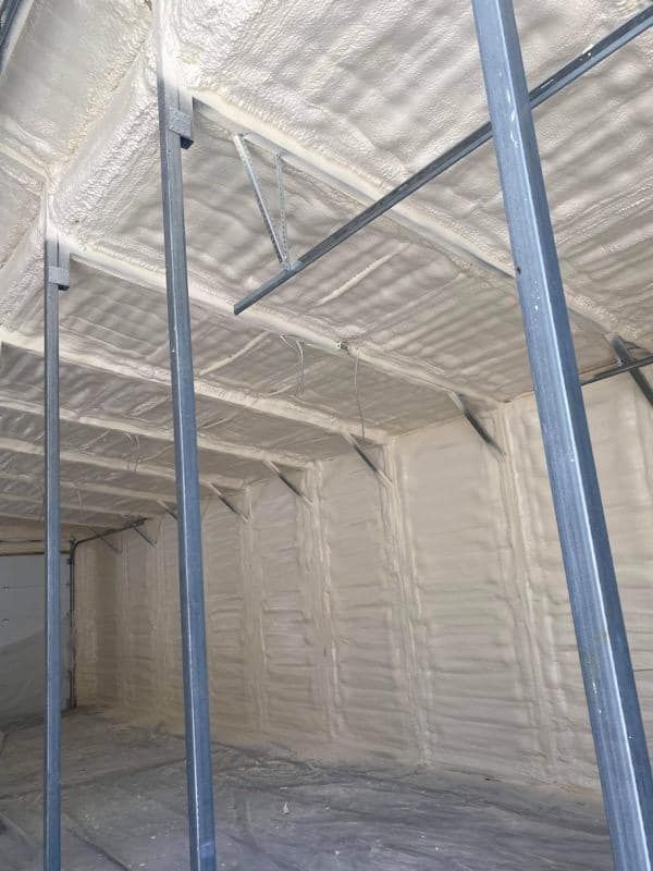 Interior of a building with grey metal support beams and walls and ceilings covered in beige spray foam insulation.