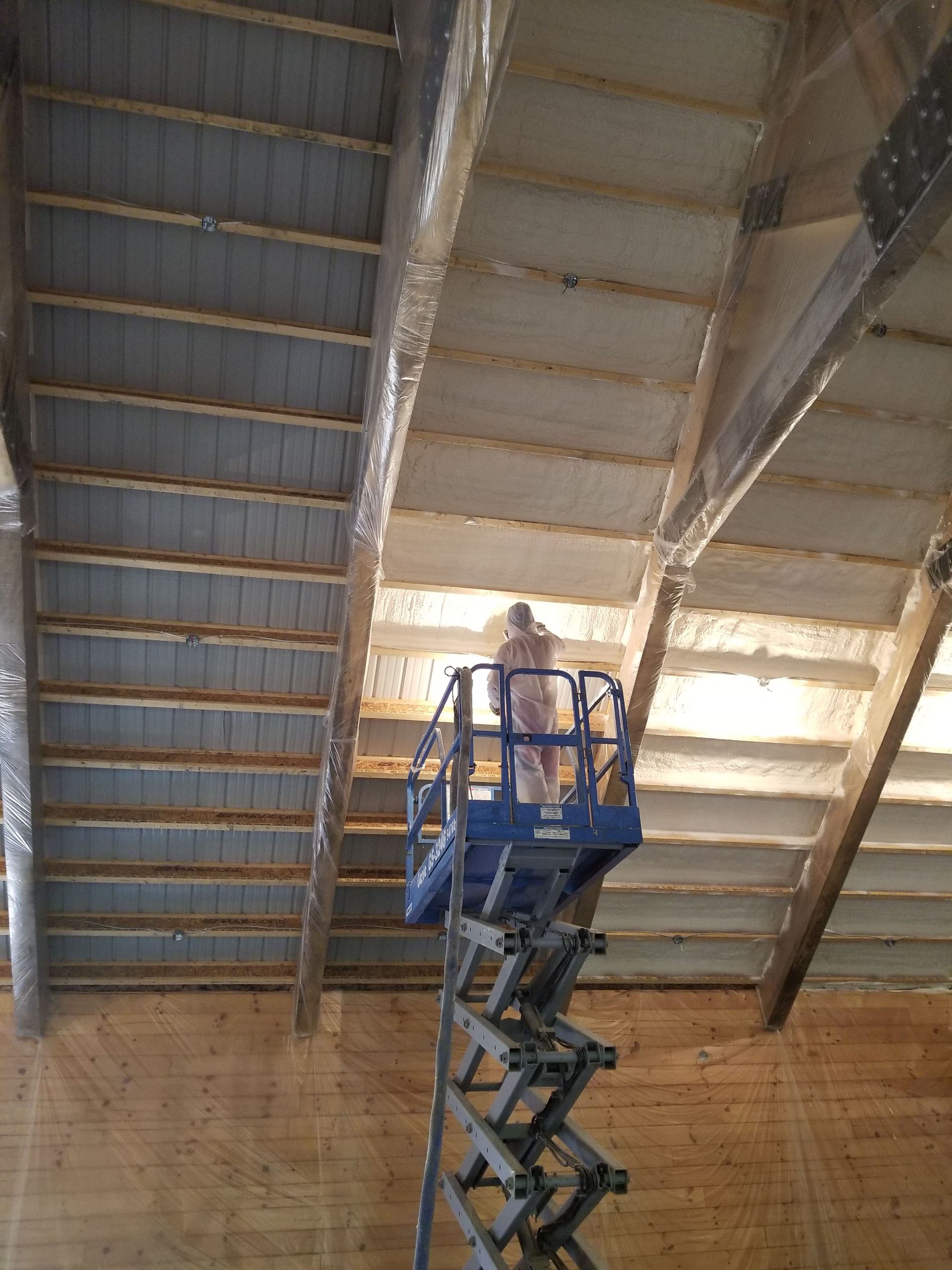 A worker in protective gear on a blue scissor lift sprays insulation onto the ceiling of a large, unfinished building.