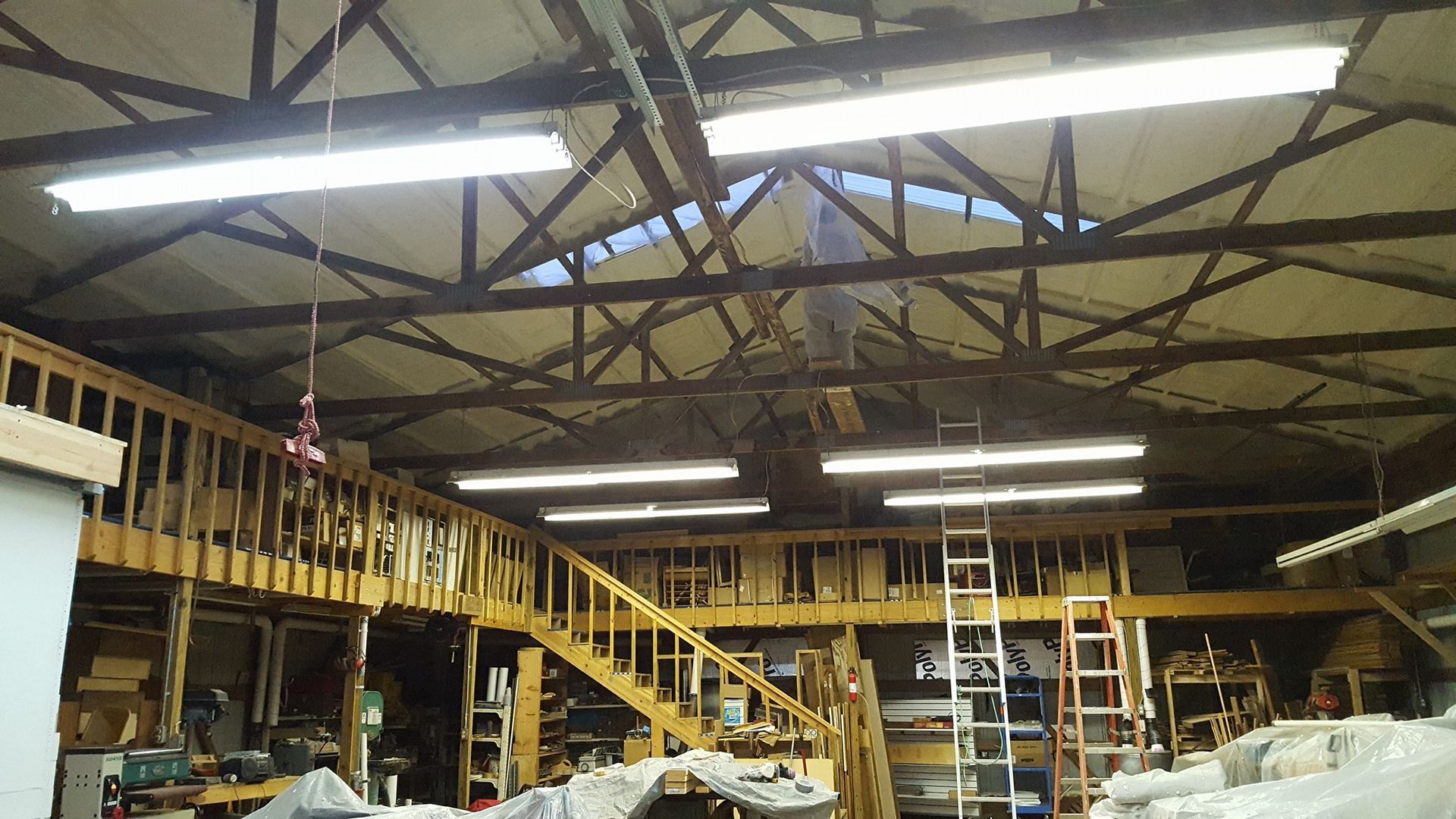 A wide-angle shot of a cluttered workshop interior with a wooden loft, stairs, and bright fluorescent ceiling lights.