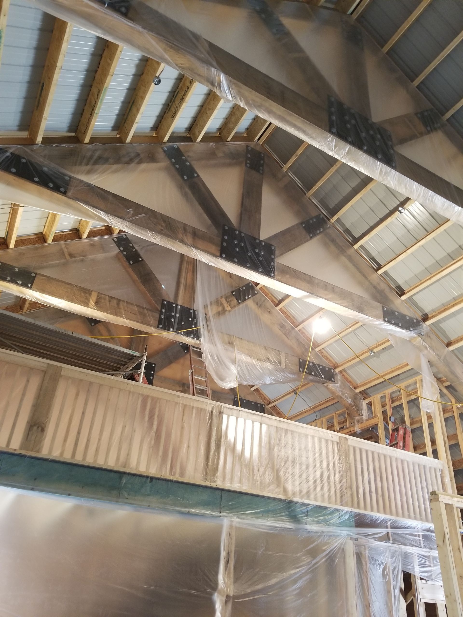 An indoor construction site showing unfinished timber roof trusses, metal bracing plates, and plastic sheeting.
