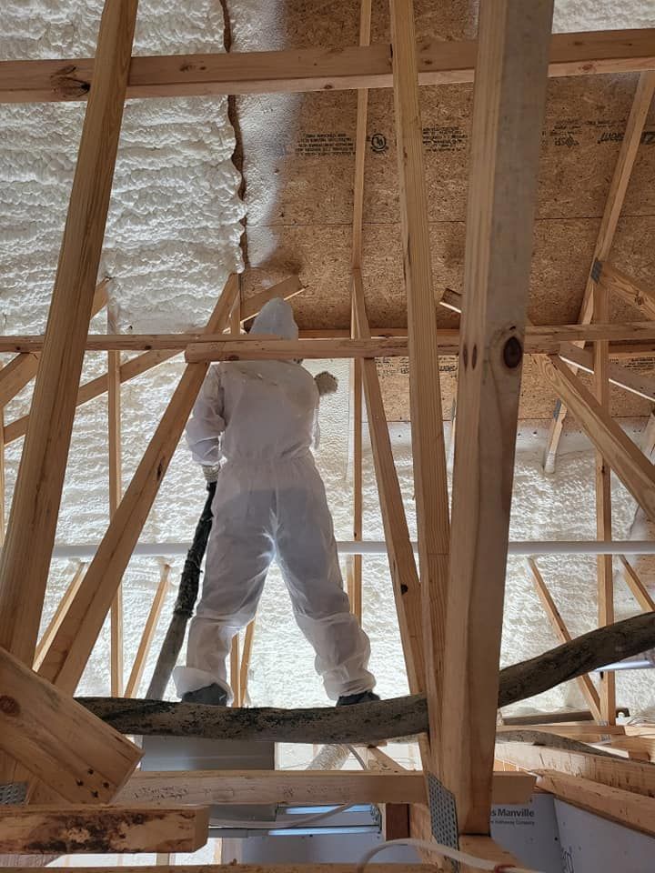 A person in protective coveralls spray-foams insulation onto the wooden roof rafters inside an unfinished attic.
