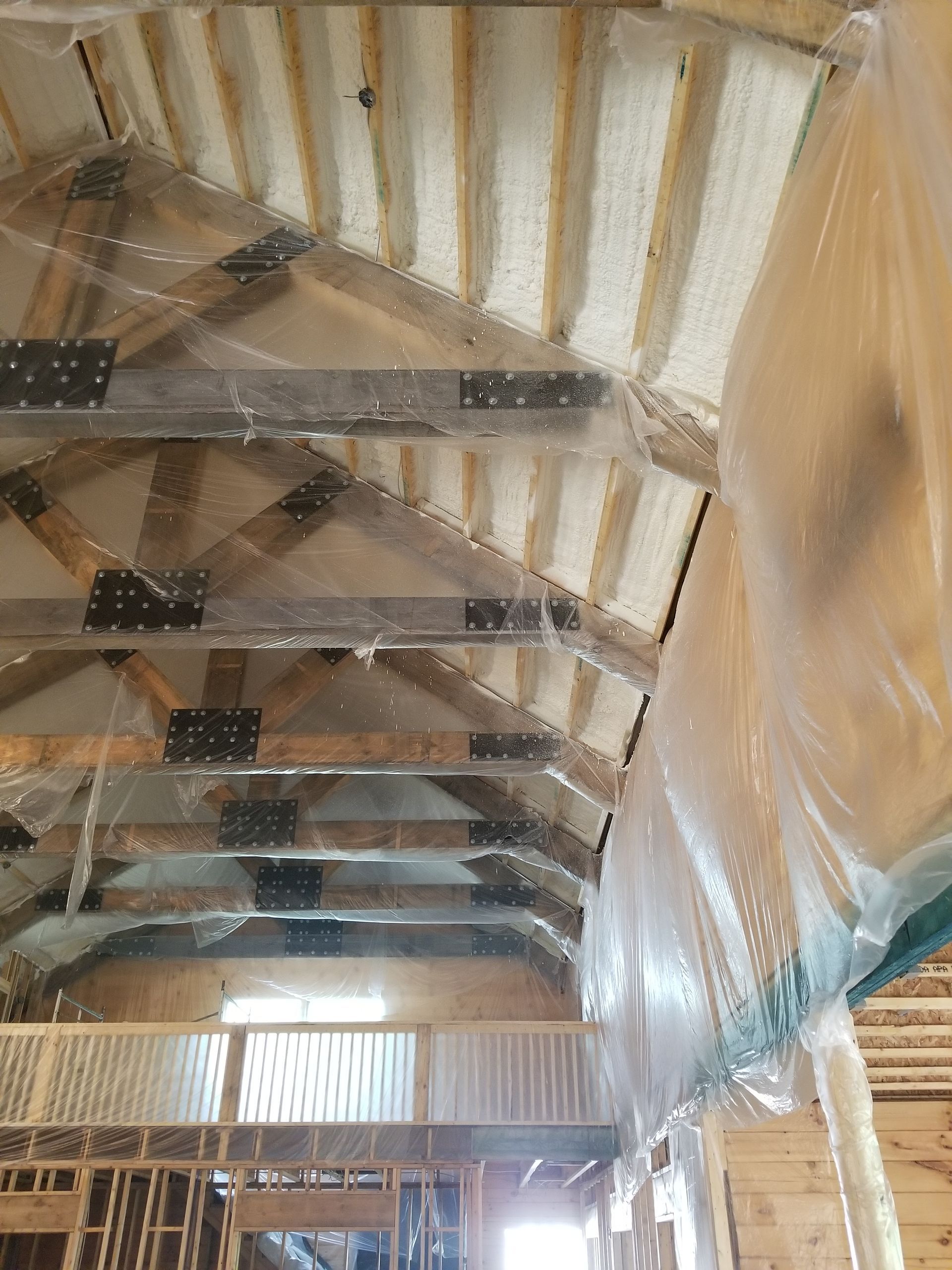 An unfinished interior space showing wooden roof trusses with spray foam insulation between them and clear plastic sheeting.