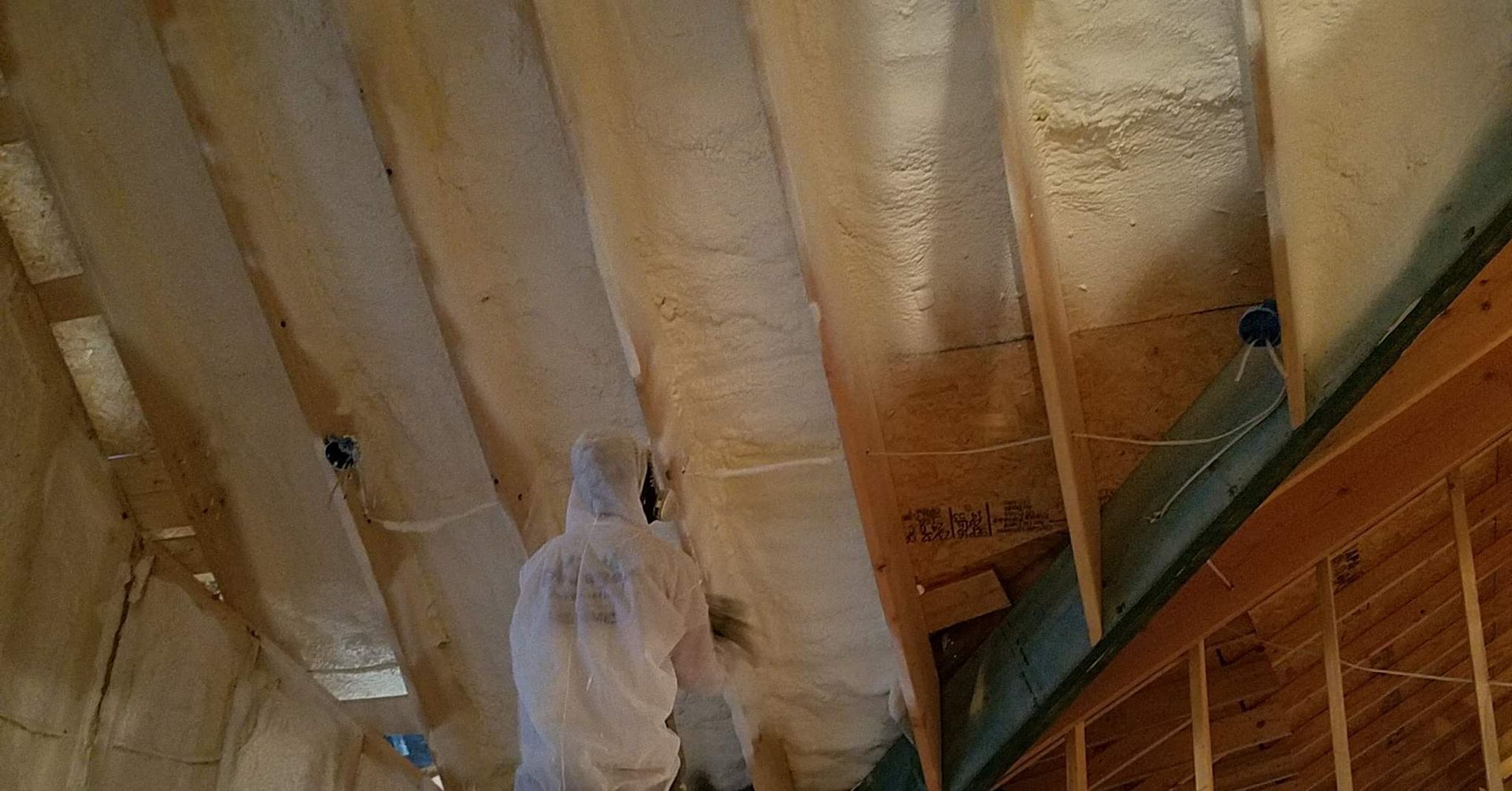 A worker in protective gear applies spray foam insulation between ceiling joists in an unfinished interior.