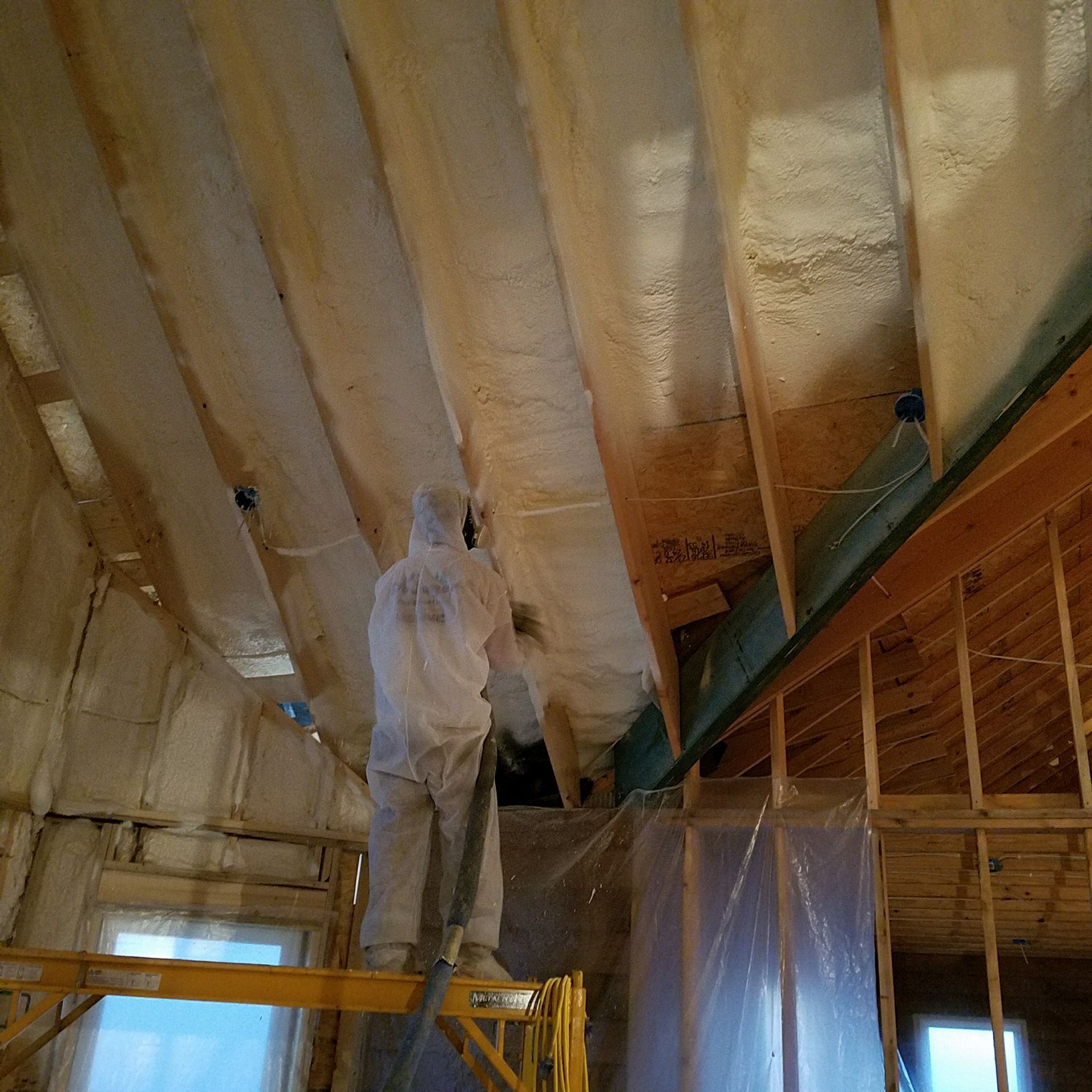 A person in a white protective suit sprays foam insulation into the rafters of an unfinished building interior.