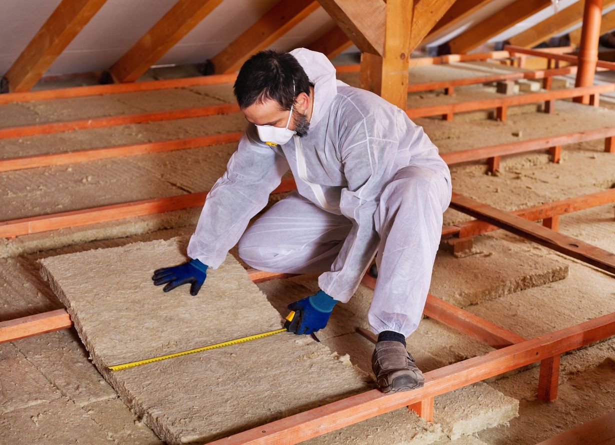A person in a protective suit and mask measures a piece of insulation in an attic.