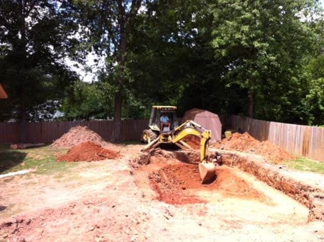 A yellow excavator is digging a hole in the dirt in a backyard.