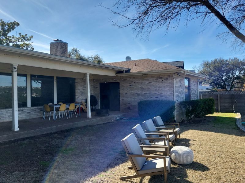 Backyard with patio, brick house, lawn, chairs, and a sunny sky.