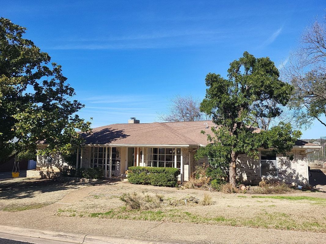 Single-story beige house with a brown roof and overgrown yard under a blue sky.