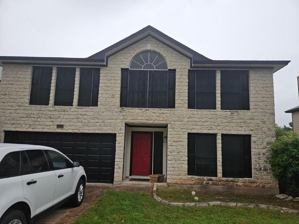 Two-story house with tan brick exterior and black shutters. Red front door and white car parked in front.