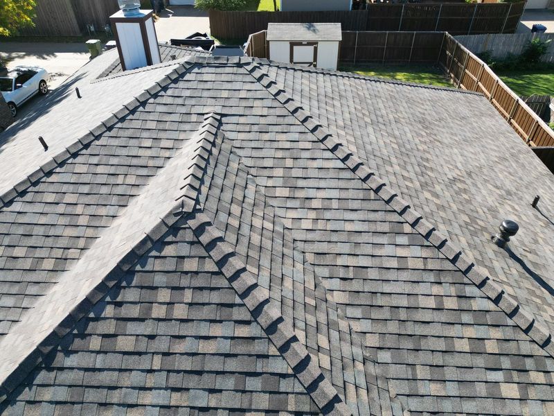 Gray and brown asphalt shingle roof on a house with a chimney and surrounding trees and fence.