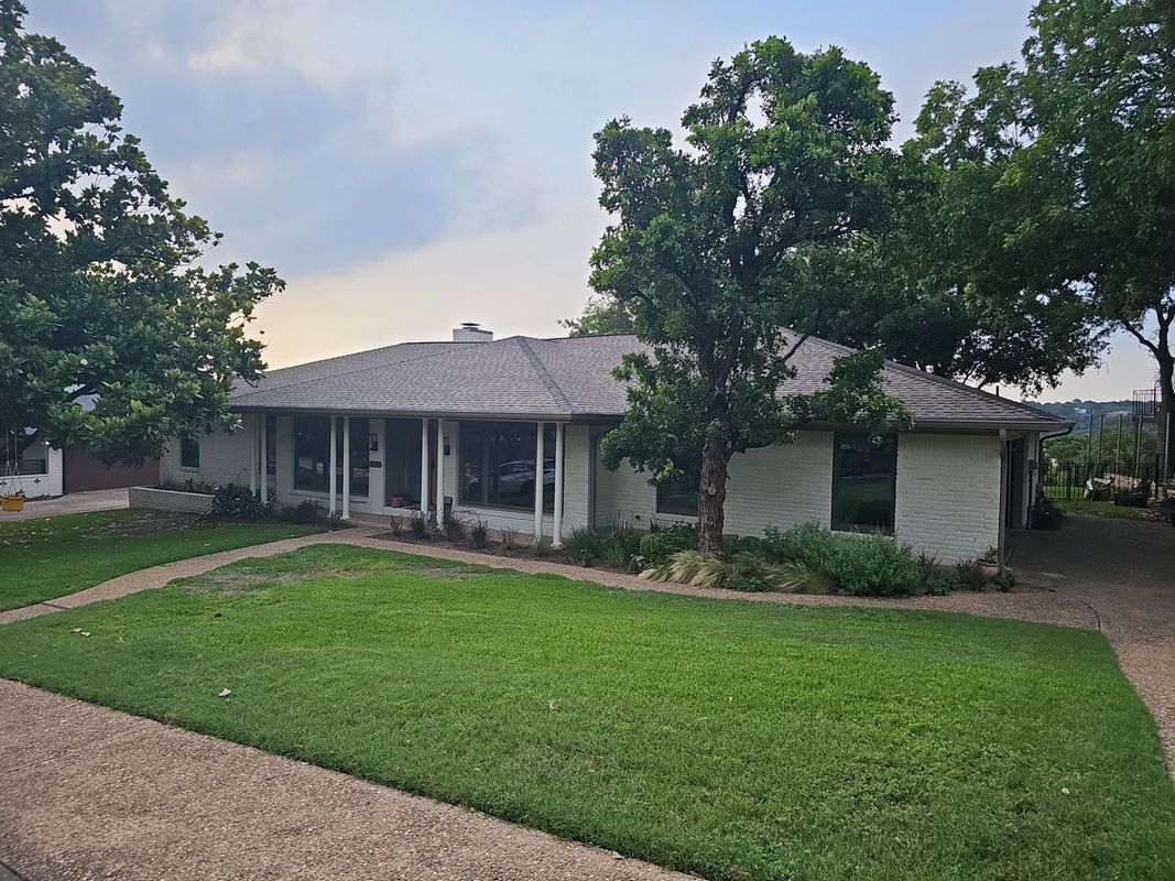 Ranch-style house with light-colored stucco exterior, surrounded by green lawn and trees.
