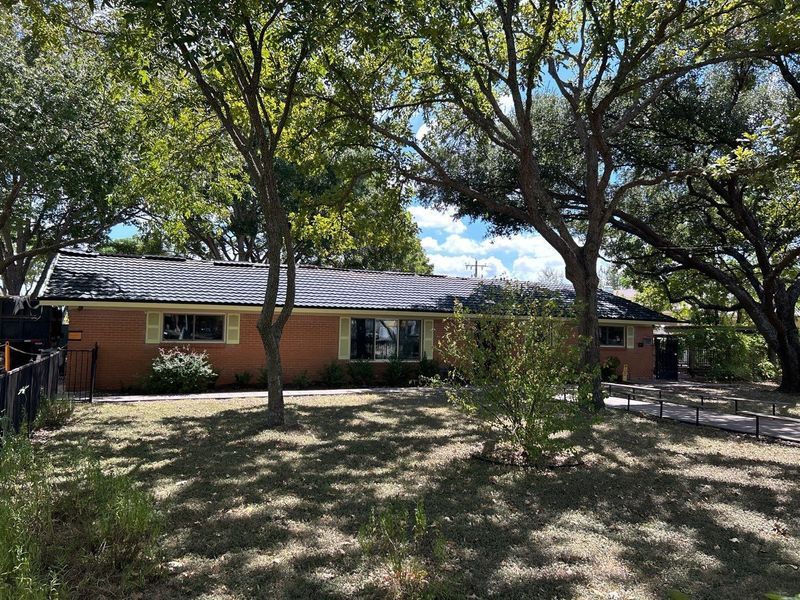 A brick ranch-style house with a dark tiled roof, surrounded by trees and a grassy lawn.