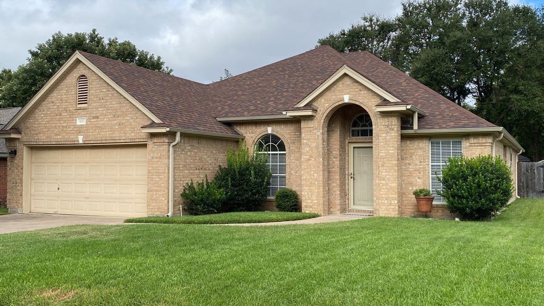 Tan brick house with brown roof and garage, green lawn, trees in the background.