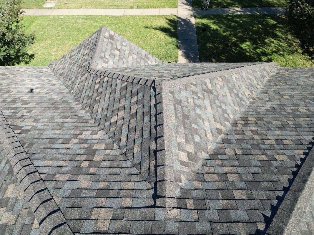 Overhead view of a house roof with dark gray and brown shingles; green grass and a sidewalk in the background.
