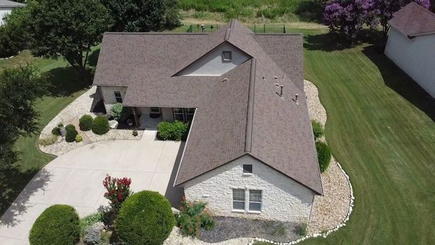 Aerial view of a house with stone siding and brown roof, situated on a green lawn with a driveway.