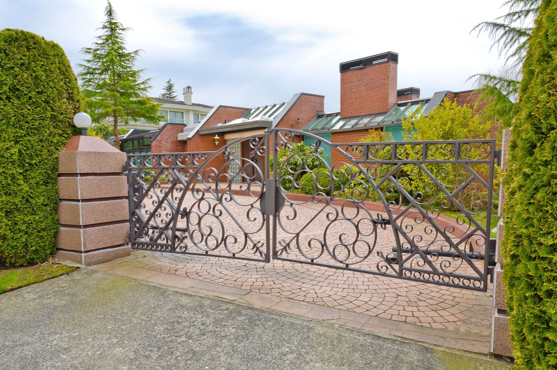 Ornate wrought-iron driveway gate in front of a brick house and hedges