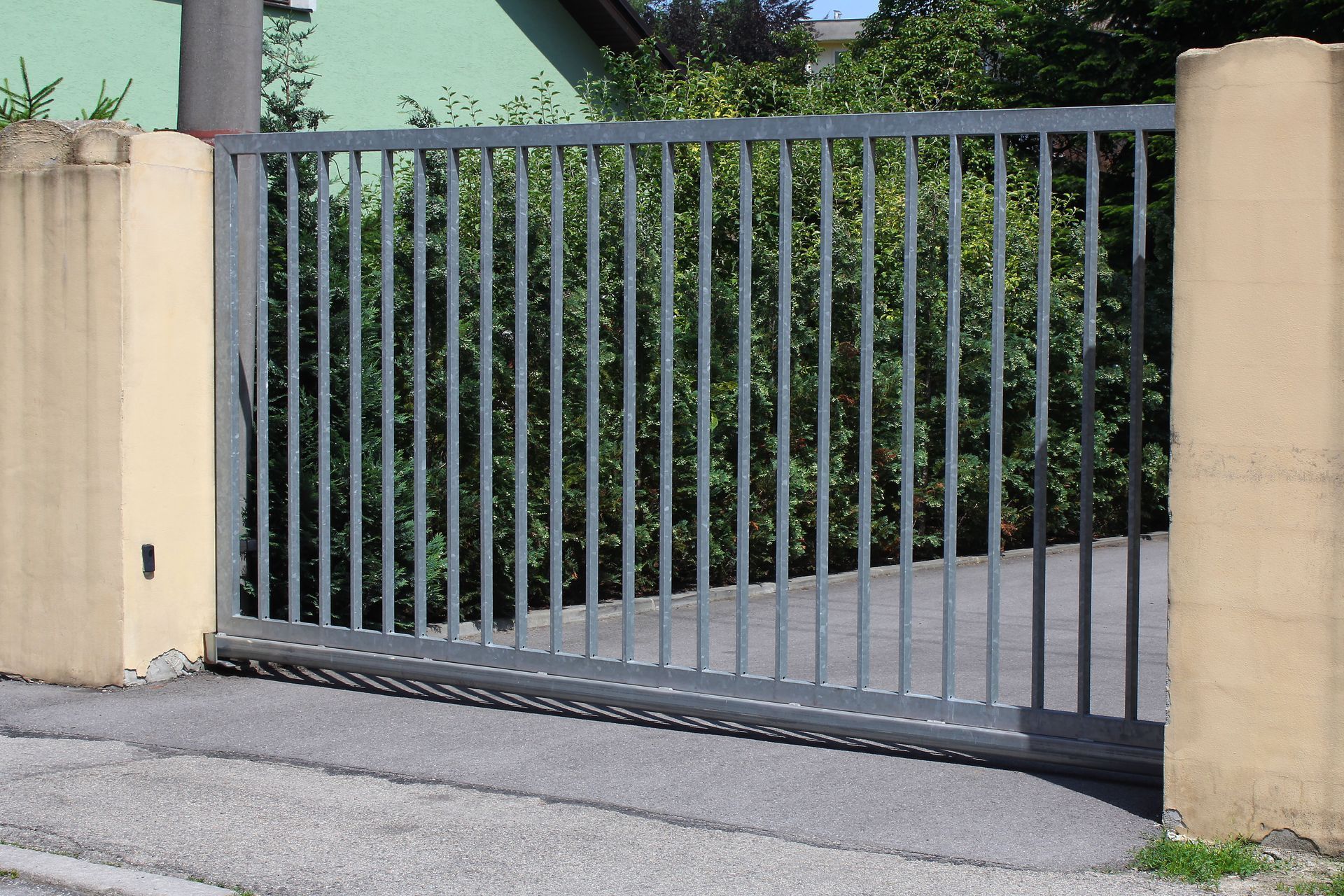 Gray metal driveway gate between beige pillars in front of dense greenery.