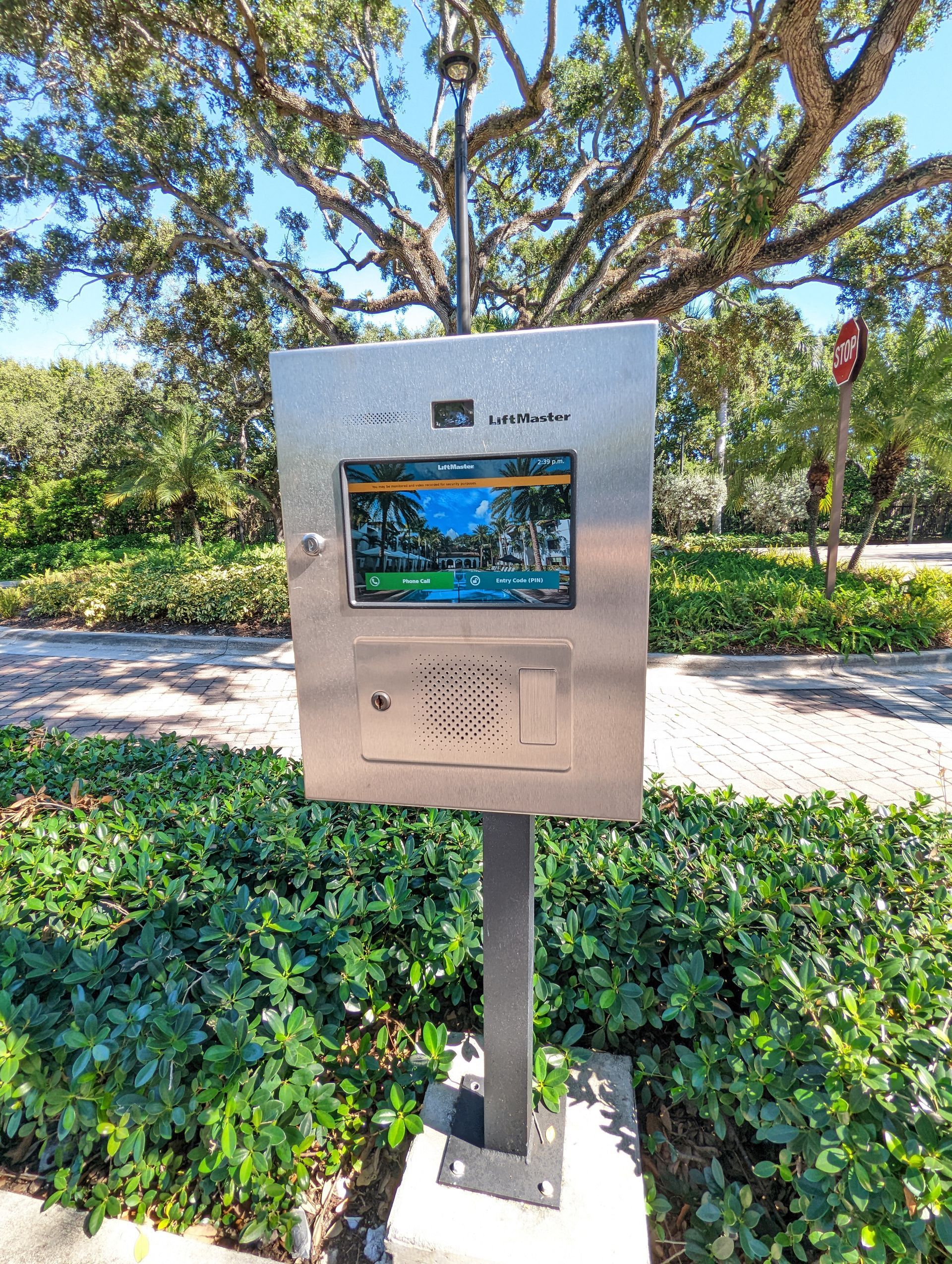 Outdoor kiosk with a small screen and speaker, mounted beside a path and greenery.